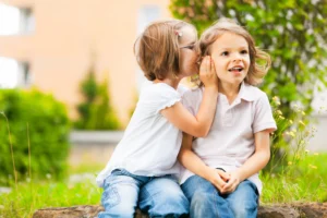 Two young children sit outside on a sunny day. One whispers in the other