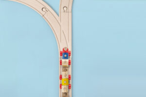 Wooden toy trains in blue, yellow, and red on a natural wood track forming a Y-shape against a light blue background, conveying playfulness.